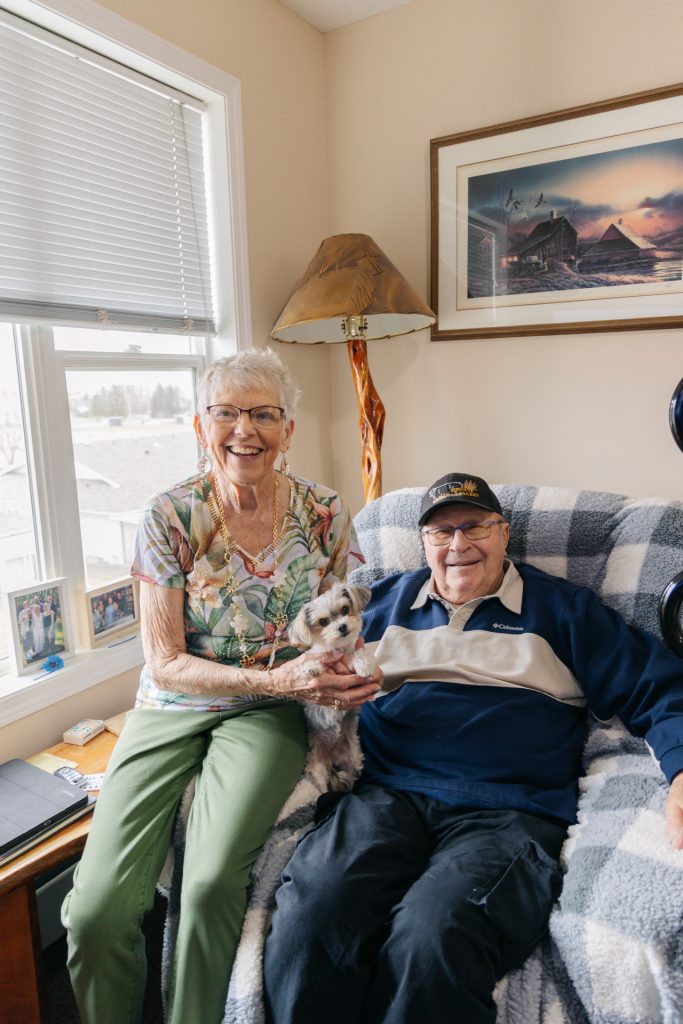 Senior couple smiling together with their small fluffy dog in their personally furnished suite at Harvest House, Olds Alberta